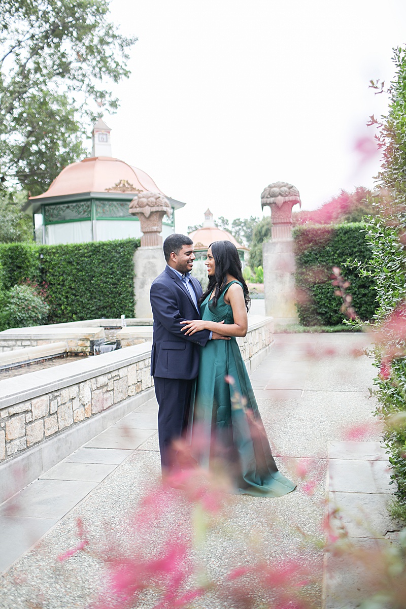 couple in navy suit and emerald green dress smile at each other through pink flowers