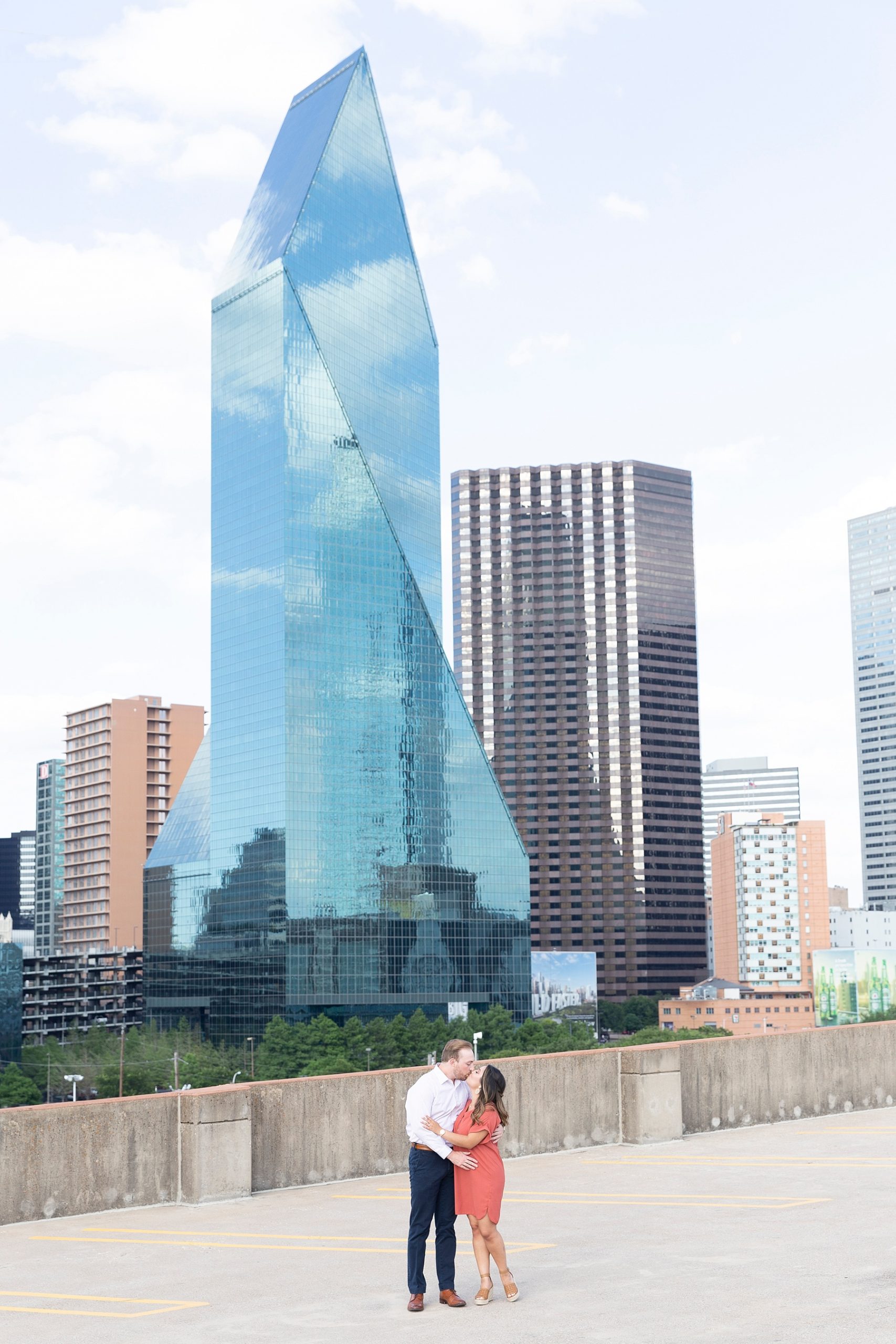 engaged couple hugs on parking garage with Dallas Skyline behind them