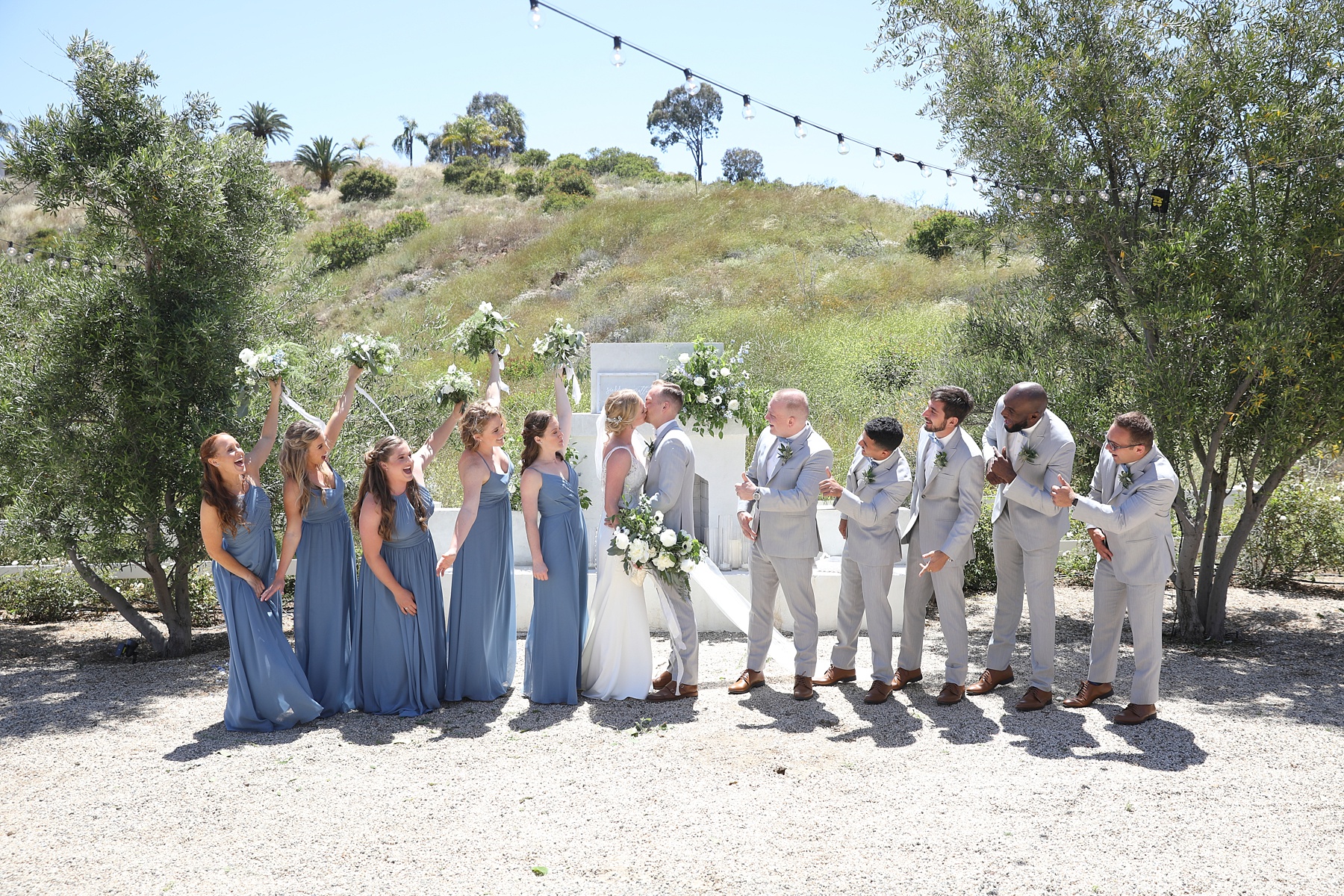 bridesmaids in blue gowns cheer while groomsmen in grey suits clap for bride and groom