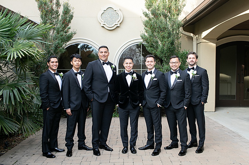 groom with 7 groomsmen in black tuxes pose outside Chapel at Ana Villa