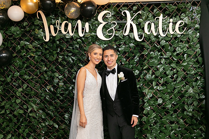 bride and groom pose in front of greenery covered wall
