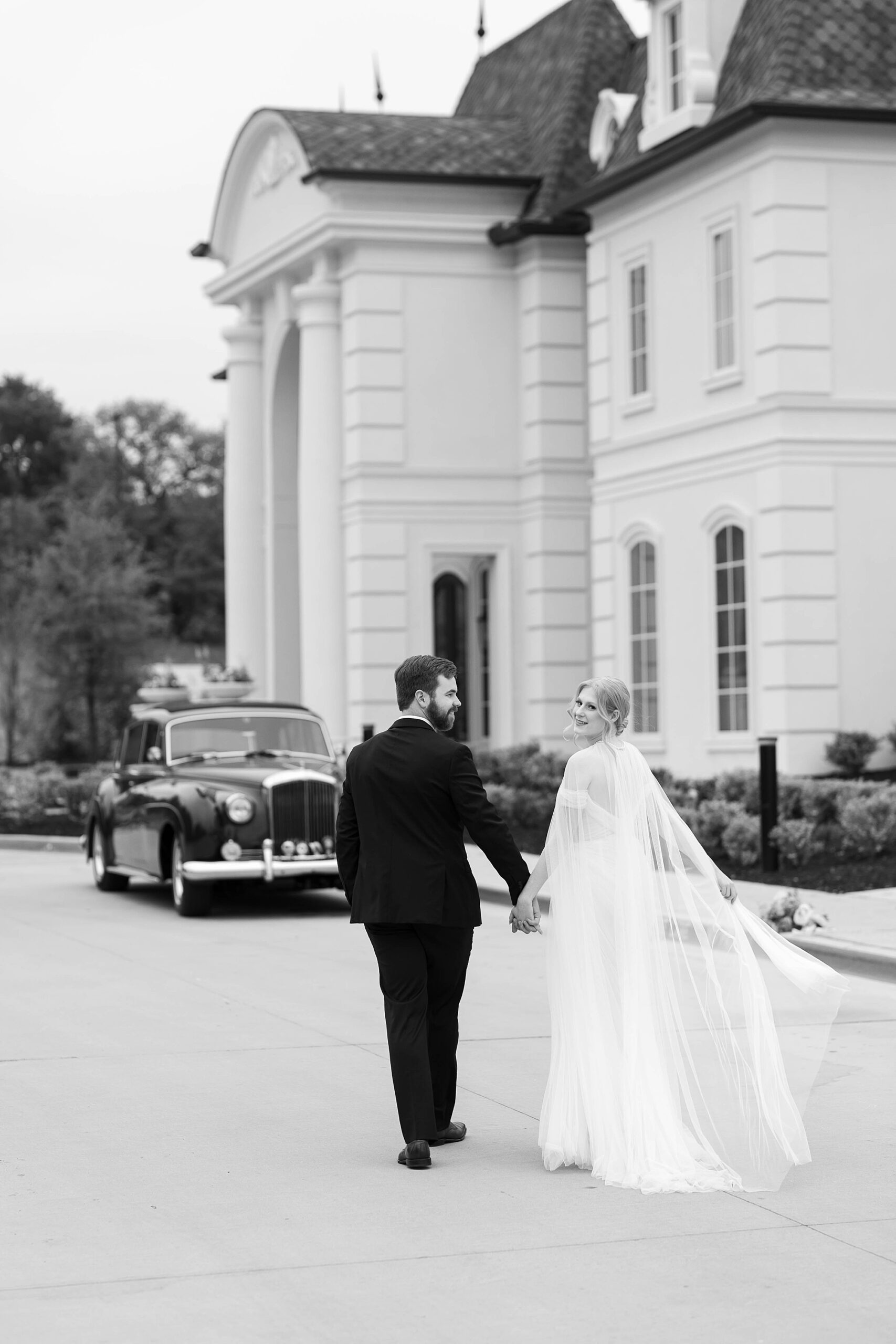 bride and groom hold hands walking together outside the Hillside Estate towards black classic car 