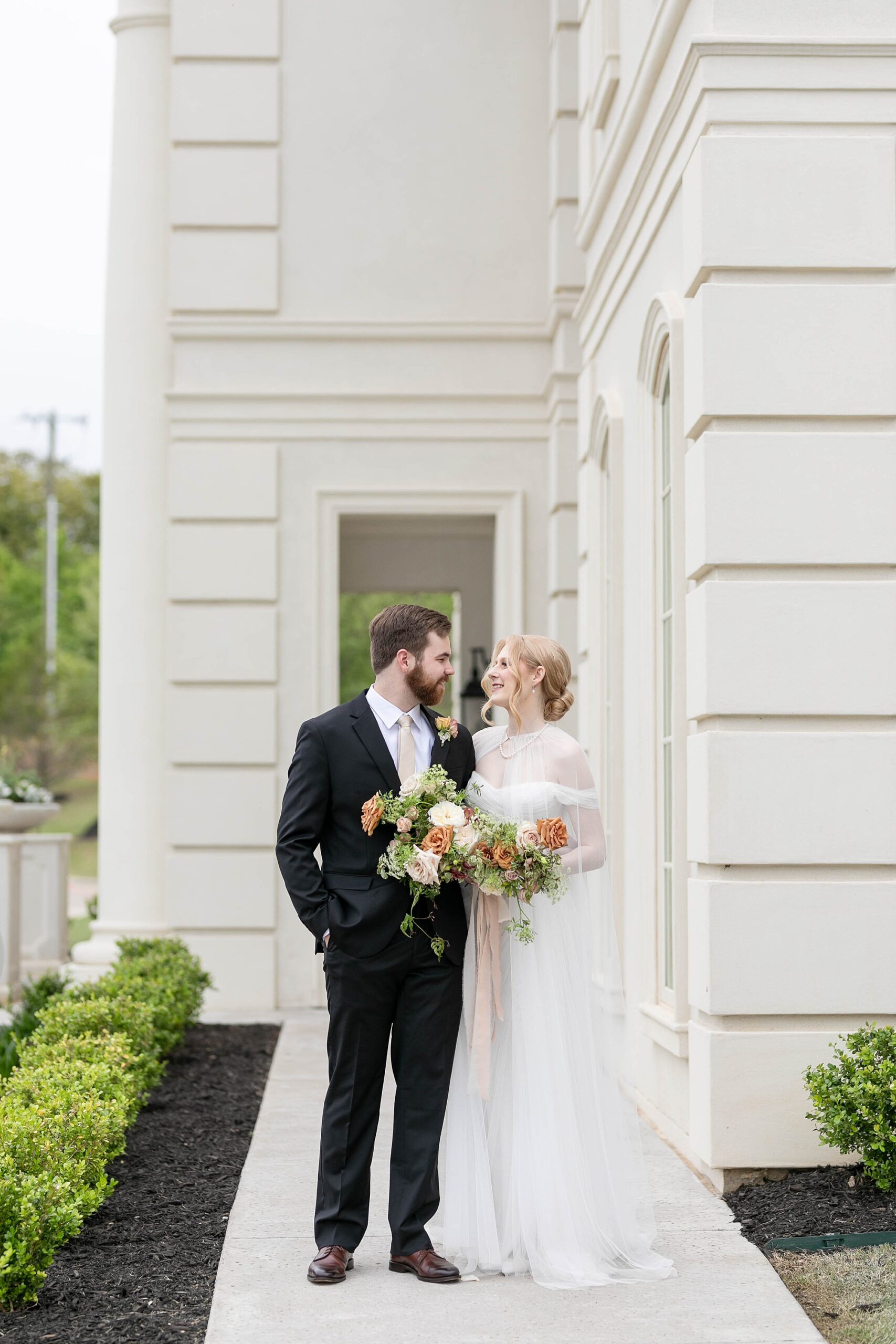 bride and groom smile at each other in front of tan pillars outside the Hillside Estate