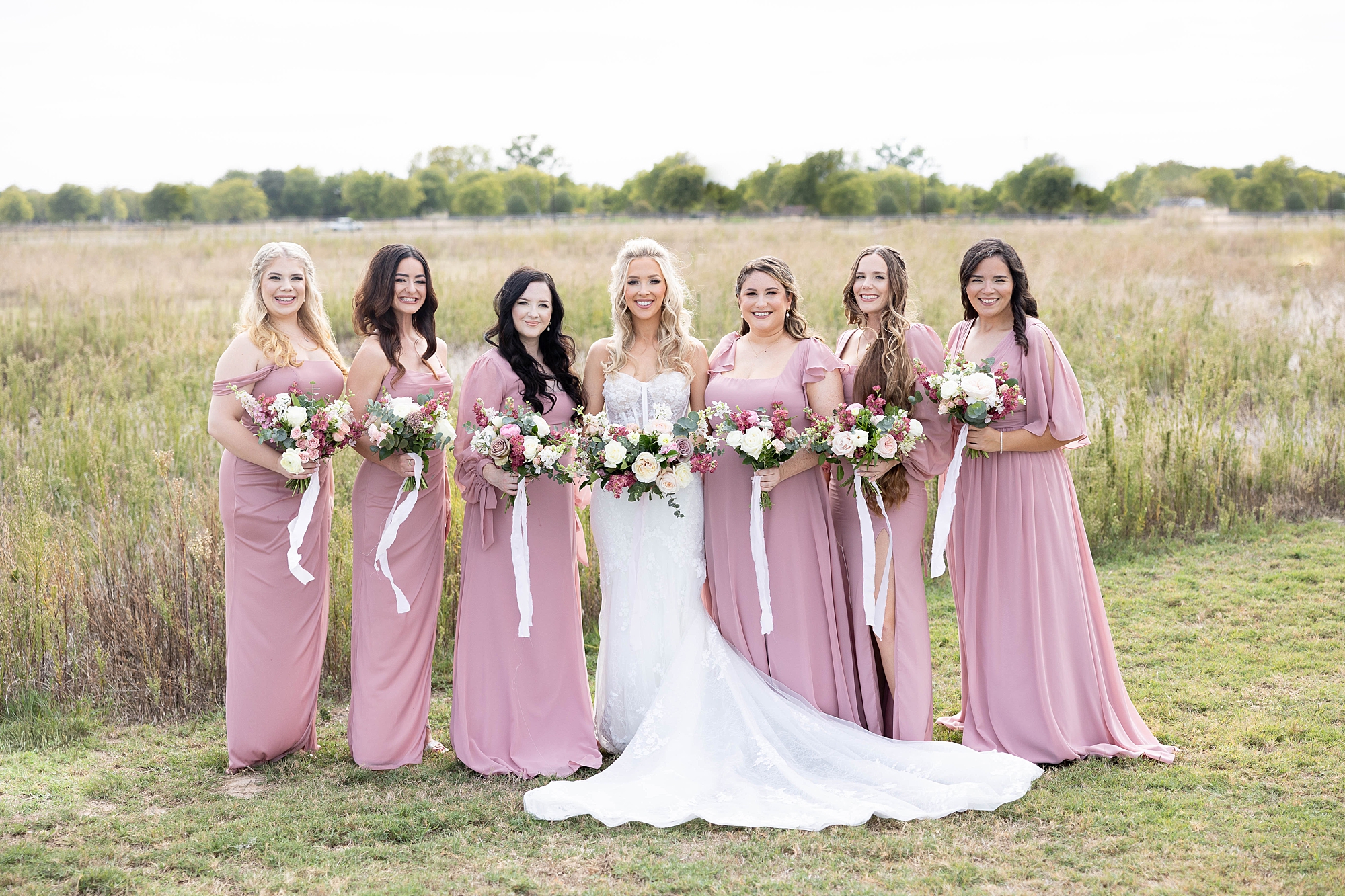 bride stands with bridesmaids in pink gowns with bouquets with white ribbons 