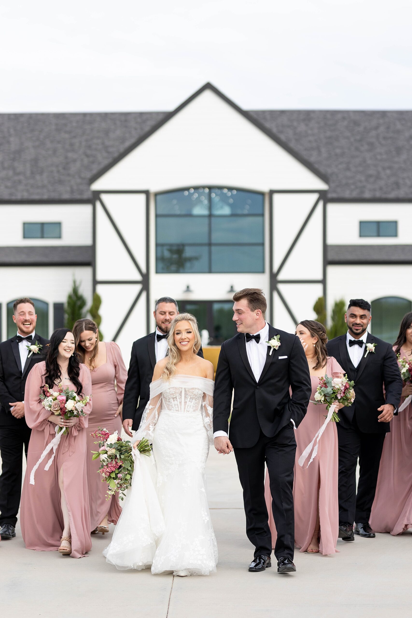 bride and groom hold hands walking in front of the Gardenia with wedding party in pink dresses and black tuxes