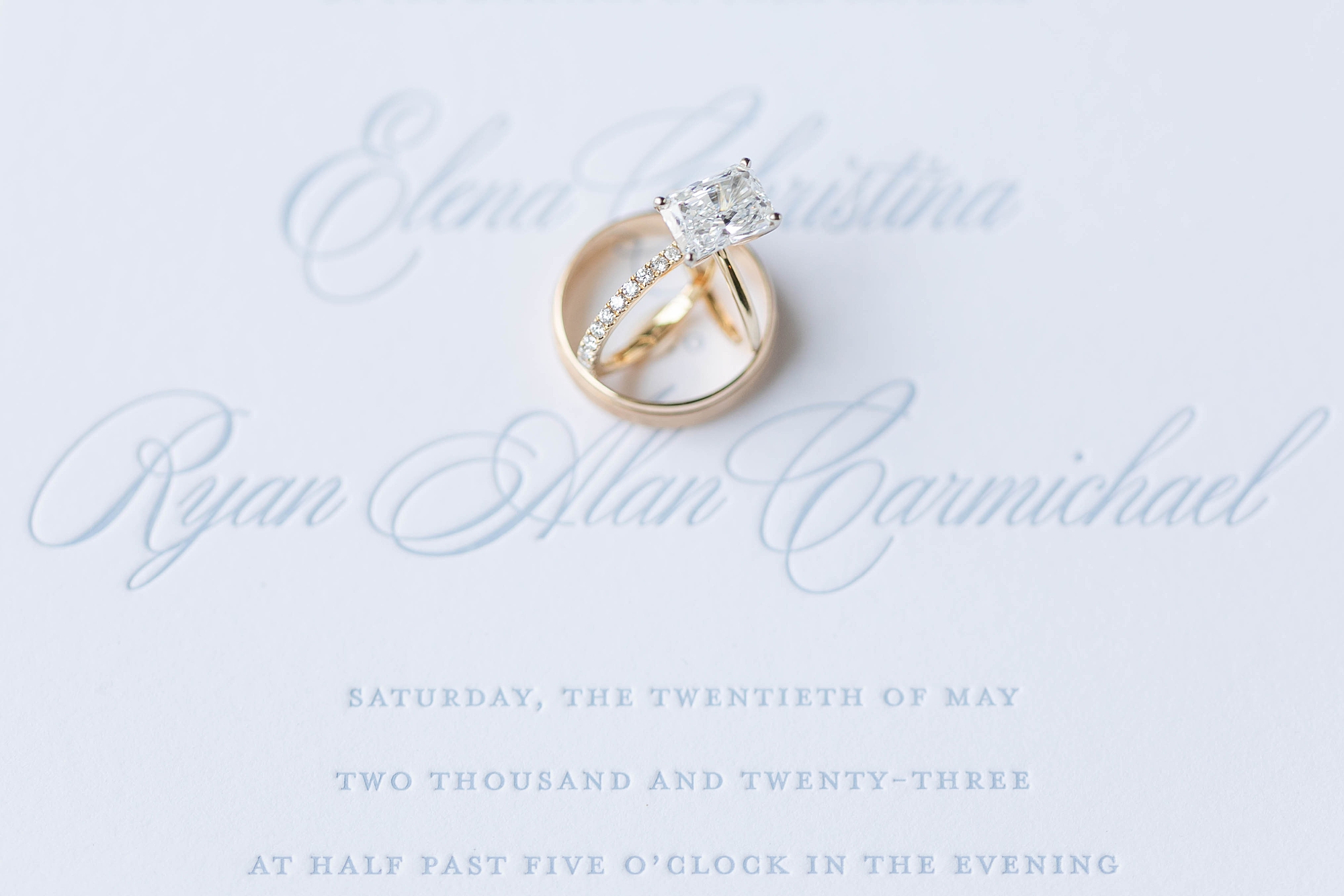 gold wedding rings rest on invitation with blue lettering