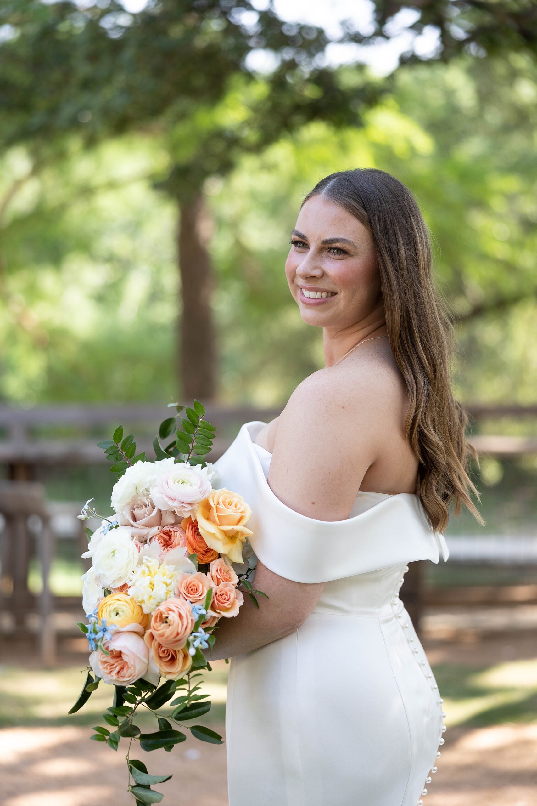 bride smiles over shoulder in off-the-shoulder wedding dress holding bouquet of peach and pink flowers 