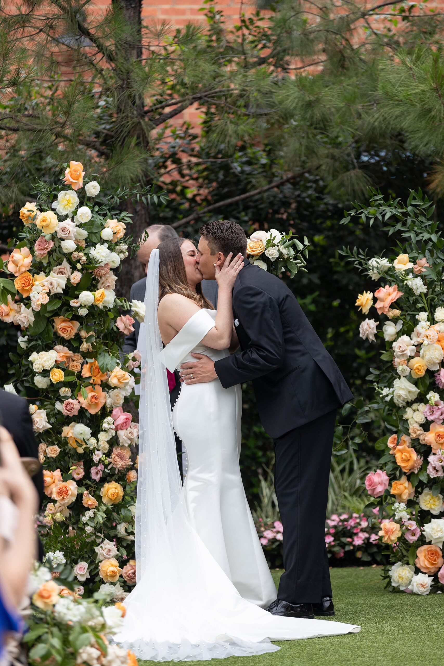 newlyweds kiss between floral arrangements of pink and peach flowers 