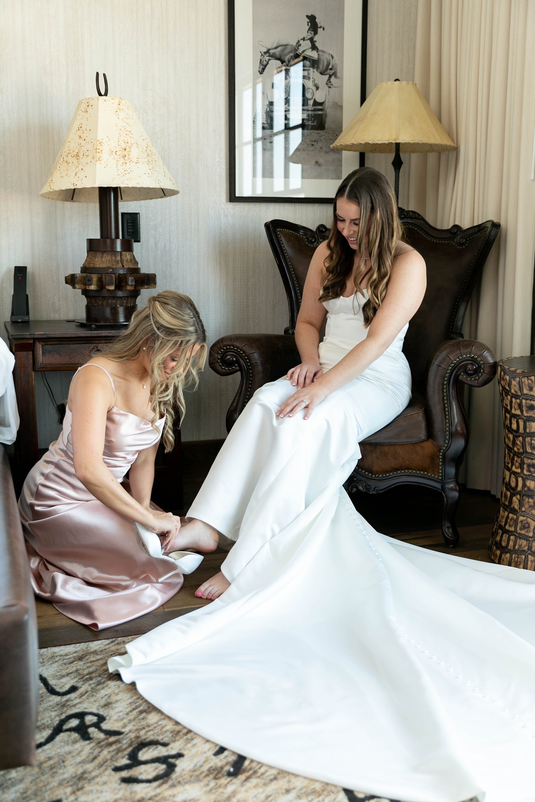 bridesmaid in metallic pink gown helps bride with shoes 