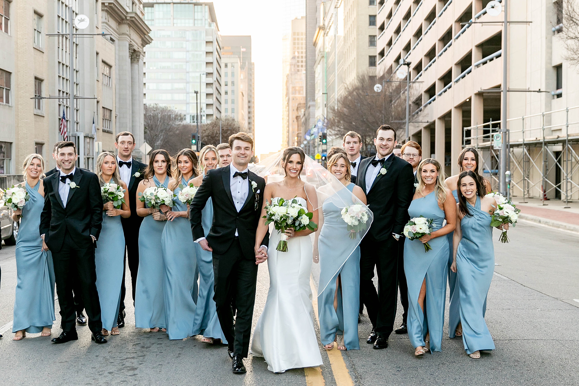 bride and groom stand with wedding party in light blue gowns and black tuxes 