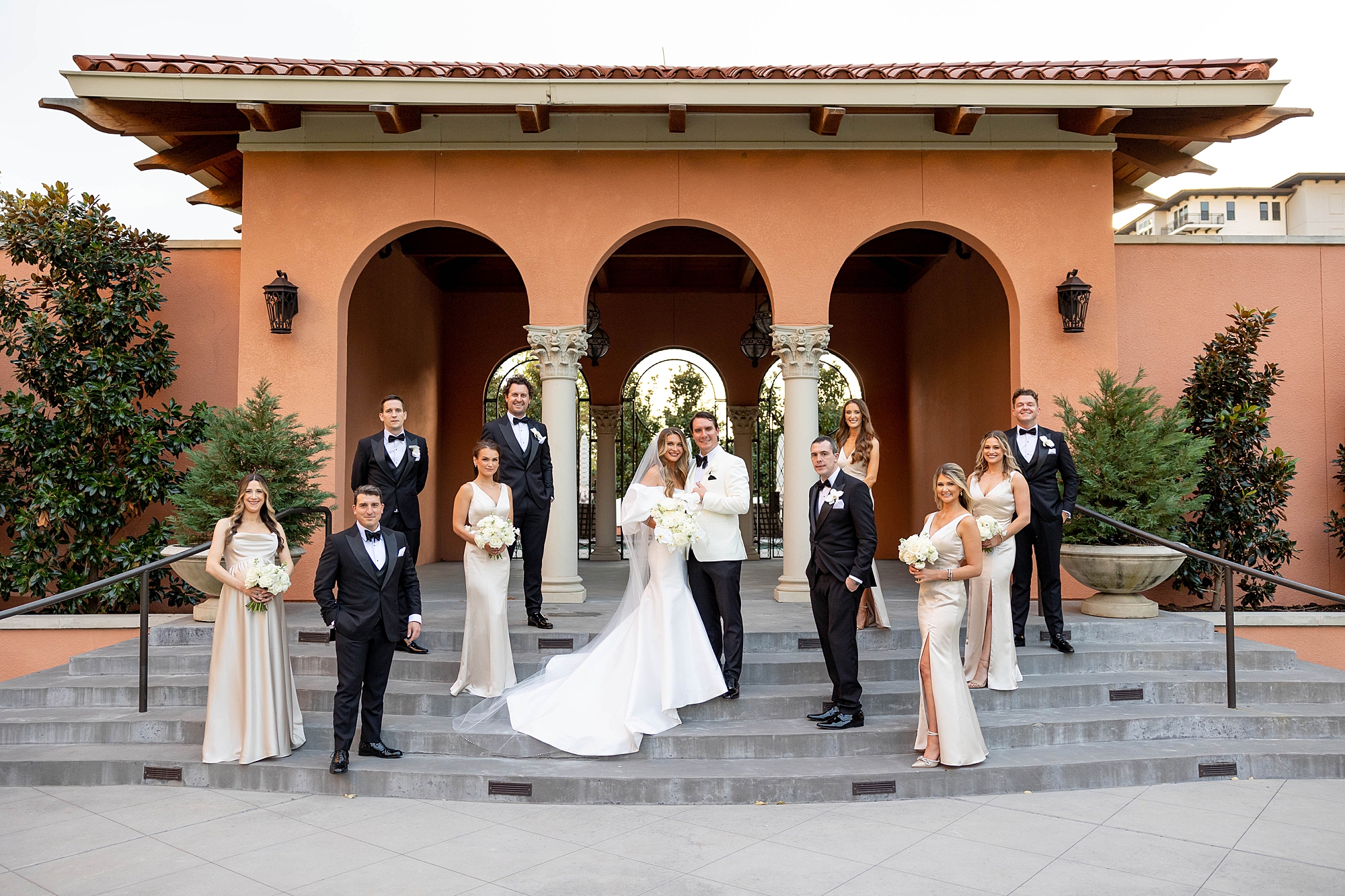bride stands on steps hugging groom with wedding party in front of archways at Rosewood Mansion