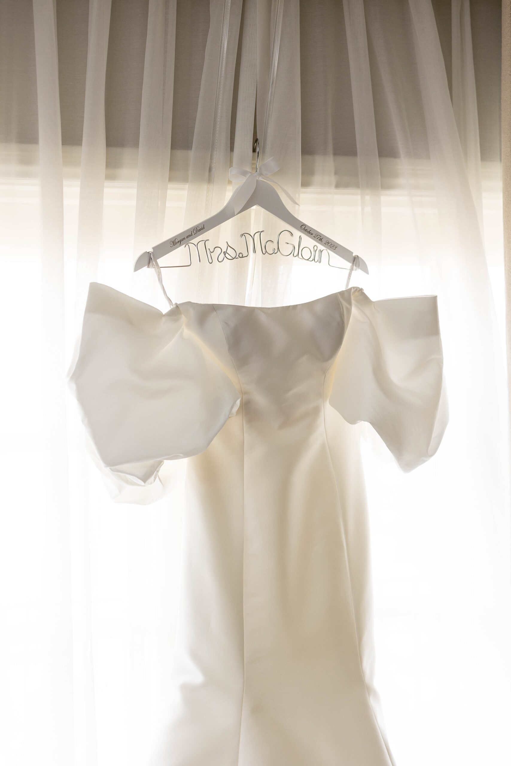 off-the-shoulder wedding gown with puffy sleeves hangs in front of white curtains 