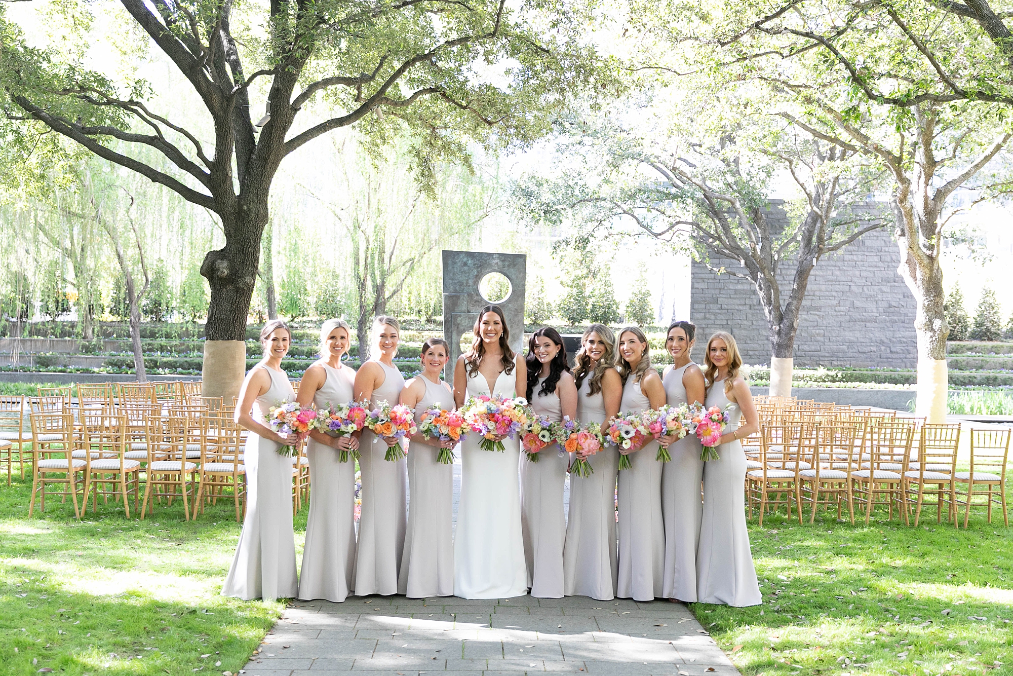 bride poses with bridesmaids in grey gowns in front of ceremony site at Nasher Sculpture Center