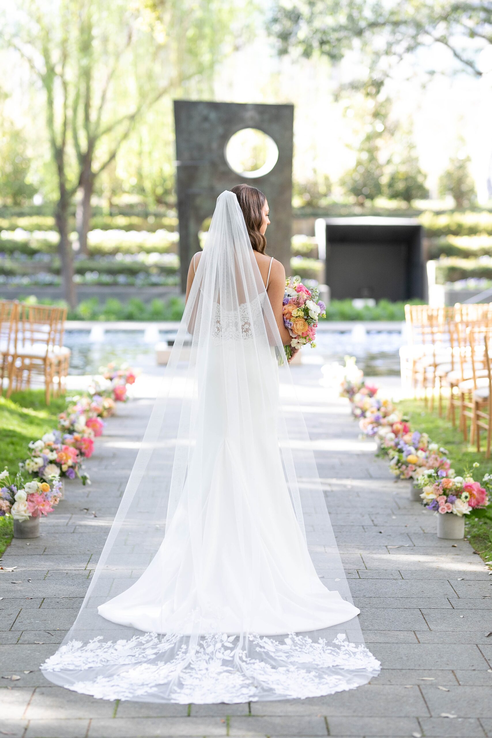 bride walks down aisle inside the Nasher Sculpture Center lined by colorful flowers 