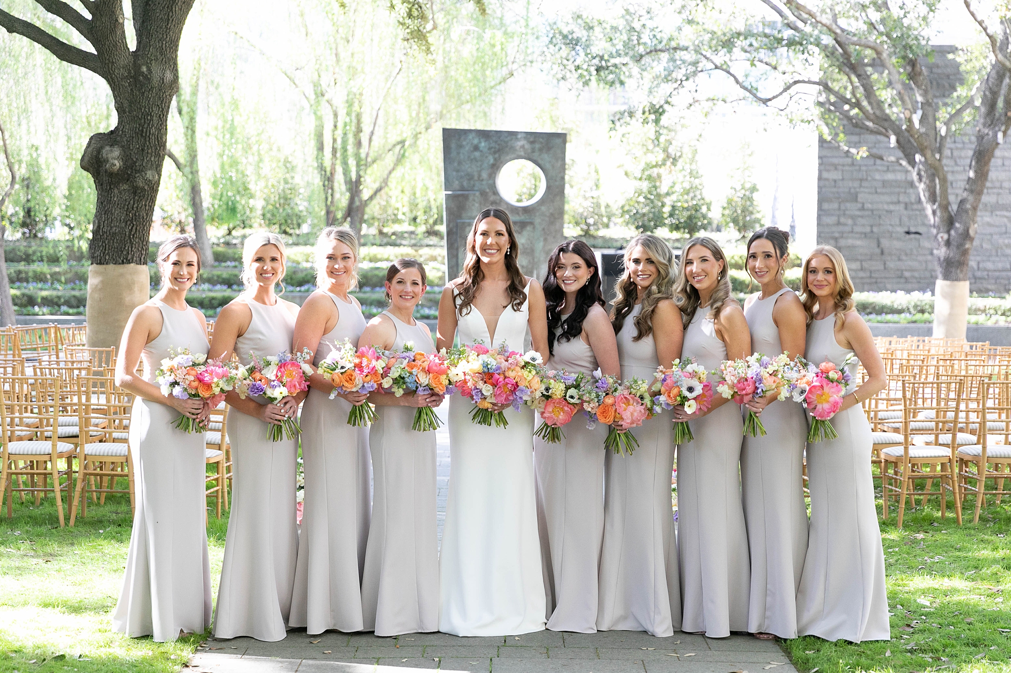 bride poses with bridesmaids in grey gowns holding bright florals for the Nasher Sculpture Center wedding
