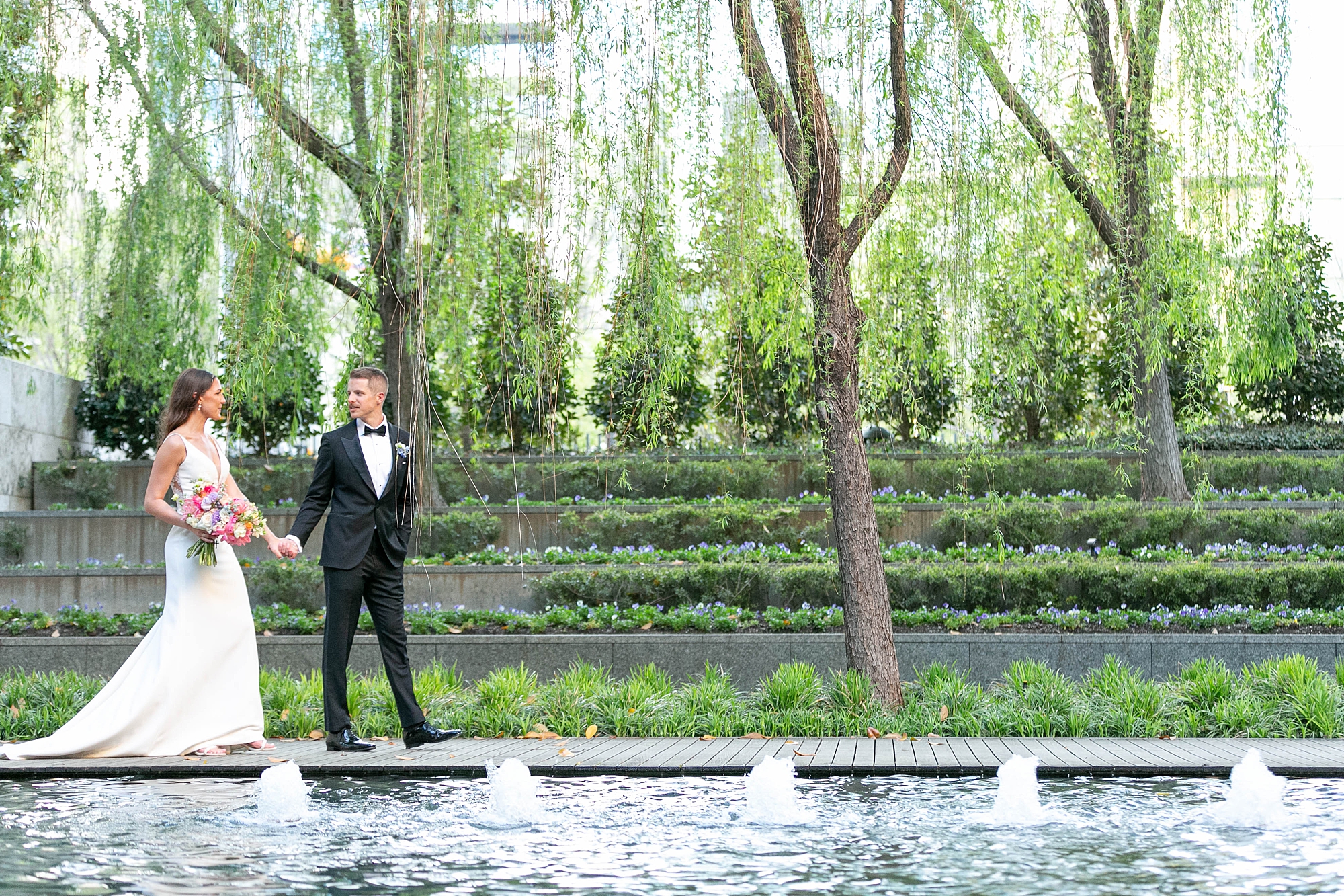 newlyweds hold hands walking along fountain inside the Nasher Sculpture Center