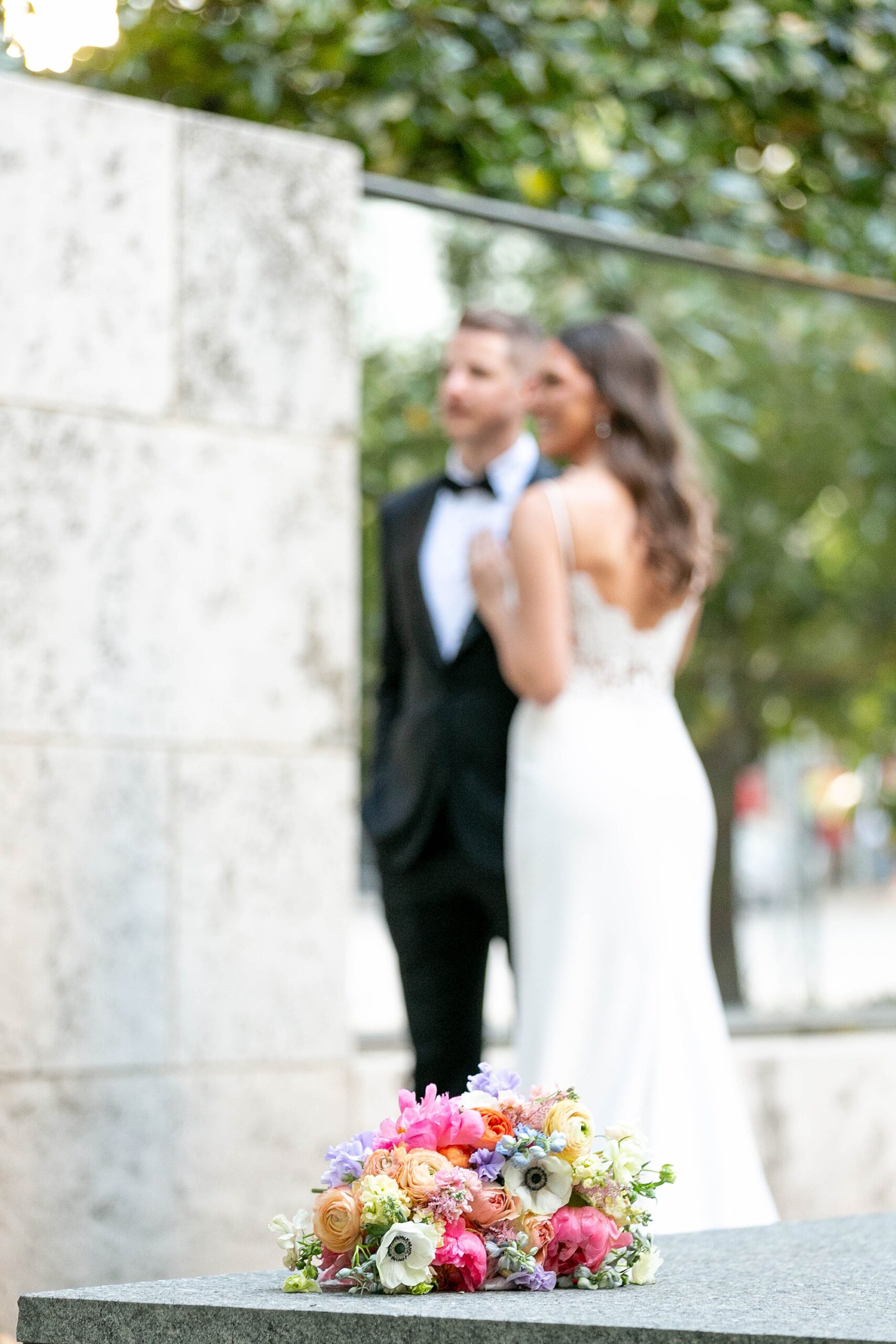 bright summer bouquet with pink and purple flowers lays at base of sculpture with newlyweds out of focus behind it 