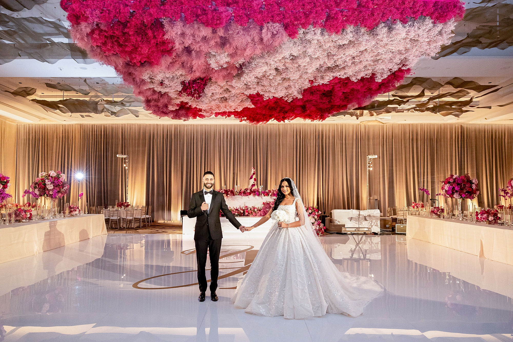 bride and groom hold hands under pink flower installation in ballroom at the Omni Frisco at the Star
